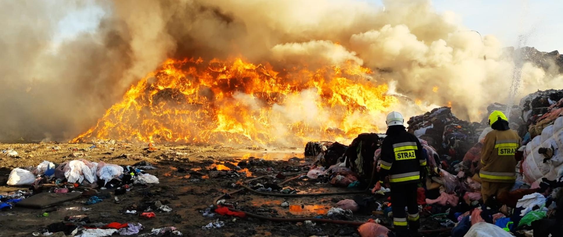 Dwaj strażacy prowadzący działania gaśnicze, Tuz obok nich porozrzucane materiały tekstylne. W tle widoczne palące się sprasowane w belach, składowane na pryzmie materiały tekstylne. Pożar miał powierzchnię 2 400m2 i kubaturę 21 600 m3, przybliżona masa materiału składowanego to 6 000 t. 