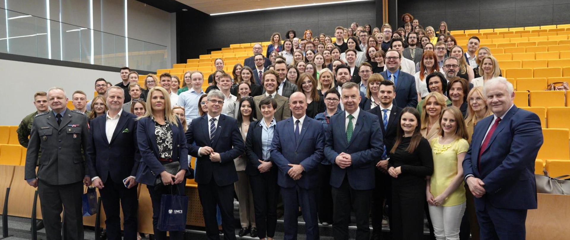 Minister Dariusz Wieczorek w otoczeniu grupy studentów. W tle aula z żółtymi krzesłami. 