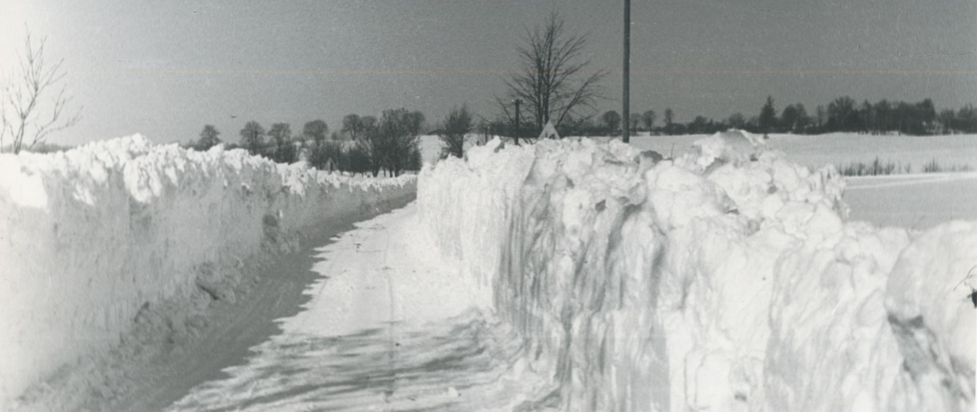 Biała droga przez pola zasypane śniegiem. Zwały śniegu odrzuconego z drogi na pobocze sięgają tak wysoko, że ledwo wystają z nich znaki drogowe. 