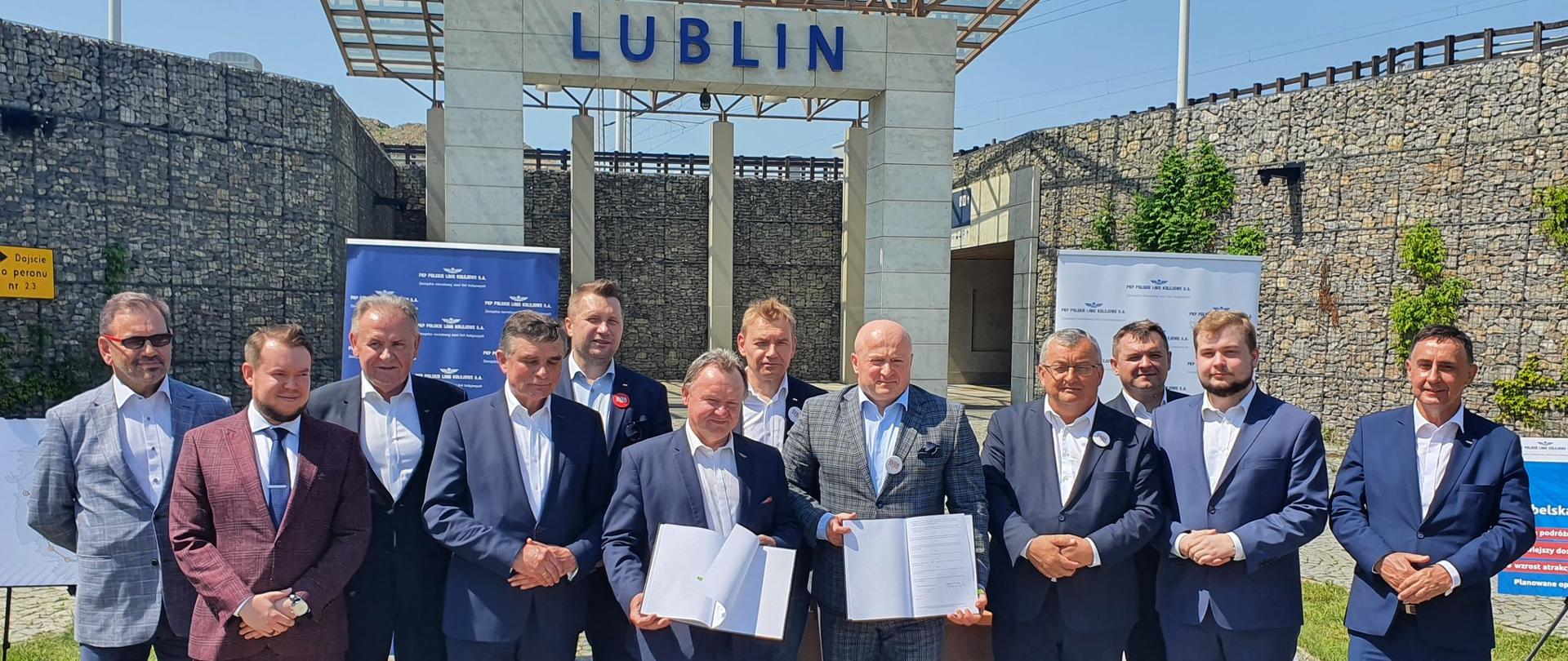 Na zdjęciu widać grupę ludzi, który pozują do zdjęcia. Panowie są ubrani w ciemne garnitury, część z nich trzyma w rękach dokument. 