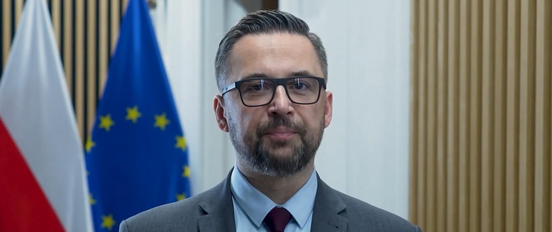 Minister Kulasek stoi i mówi na tle ściany z pionowych drewnianych listewek, za nim po lewej flagi Polski i UE.