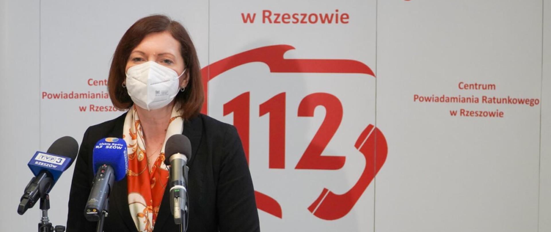 Wojewoda podkarpacki Ewa Leniart zabiera głos podczas konferencji prasowej z okazji Europejskiego Dnia Numeru Alarmowego 112. Stoi na tle banneru z czerwonymi napisami na białym tle: Centrum Powiadamiania Ratunkowego w Rzeszowie.