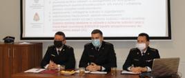 Na sali narad przy stole prezydialnym prowadzący spotkanie. Widoczni są: starszy brygadier Grzegorz Wilczyński – Zastępca Komendanta Miejskiego PSP w Łomży, brygadier Adam Kalinowski – Naczelnik Wydziału Operacyjnego KM PSP w Łomży oraz starszy kapitan Paweł Leszczyński – Zastępca Naczelnika Wydziału Operacyjnego KM PSP w Łomży.