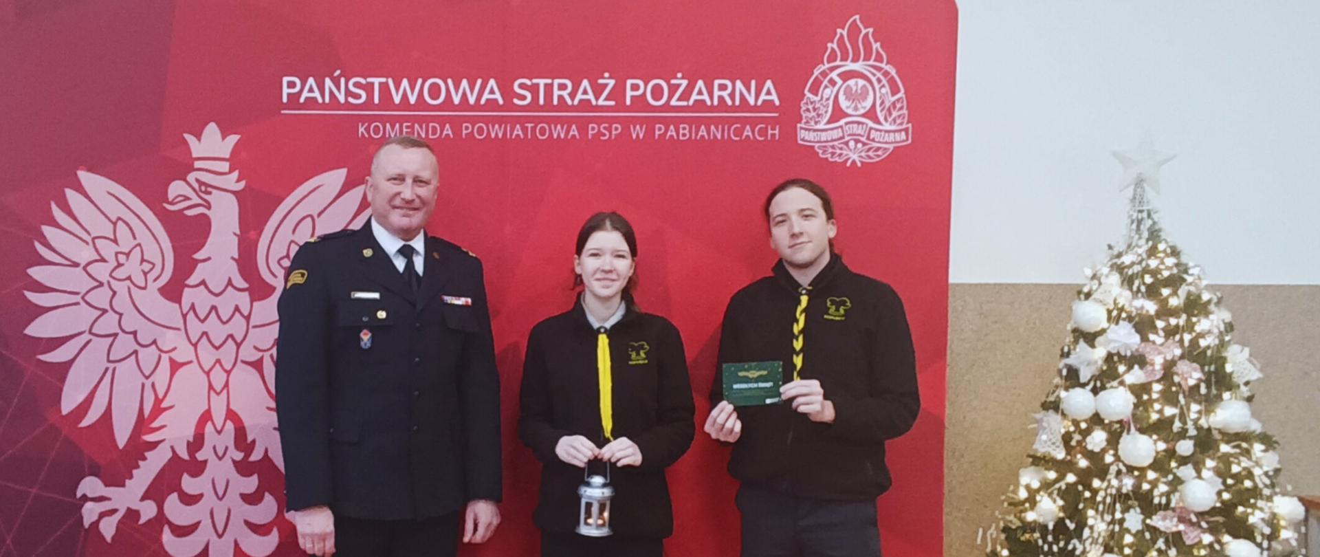 Na zdjęciu Komendant Powiatowy PSP w Pabianicach - młodszy brygadier Jacek Karcz wraz z harcerzami z Hufca w Pabianicach podczas przekazania symbolicznego Światełka Pokoju. W tle czerwona ścianka PSP, a po prawej świąteczna choinka.