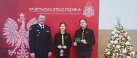 Na zdjęciu Komendant Powiatowy PSP w Pabianicach - młodszy brygadier Jacek Karcz wraz z harcerzami z Hufca w Pabianicach podczas przekazania symbolicznego Światełka Pokoju. W tle czerwona ścianka PSP, a po prawej świąteczna choinka.
