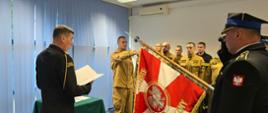 Zdjęcie przedstawia oficjalną ceremonię strażacką w pomieszczeniu biurowym, najprawdopodobniej w Polsce, o czym świadczą napisy na sztandarze i mundury.
Oto szczegółowy opis tego, co widać na zdjęciu:
• Grupa ludzi: Po prawej stronie widać grupę strażaków w piaskowych mundurach służbowych (polowych/koszarowych) i jednego w ciemnym mundurze z białymi pagonami. Wszyscy stoją na baczność.
• Oficer odczytujący dokument: W centralnej części, tyłem do obiektywu, stoi oficer w ciemnym mundurze wyjściowym lub galowym z baretkami odznaczeń. Trzyma otwarty dokument w białej oprawie i czyta jego treść, prawdopodobnie rotę przysięgi, nominację lub rozkaz.
• Strażak trzymający sztandar: Na lewo od czytającego oficera stoi strażak w piaskowym mundurze, który trzyma oburącz pochylony sztandar.
• Sztandar: Jest to biało-czerwony sztandar ze złotą frędzlą. Na czerwonym płacie widać godło Polski – orła w koronie. Nad nim widnieje napis "OJczyzny" (część słowa "Ojczyźnie"), a po bokach widać fragmenty innych napisów (prawdopodobnie "Służyć Bogu i Ojczyźnie", "Państwowa Straż Pożarna" lub nazwa jednostki, choć są one częściowo zasłonięte).
• Inni oficerowie: Po lewej stronie zdjęcia widać fragment sylwetki innego oficera w ciemnym mundurze wyjściowym z rogatywką. Na pagonach widać stopień (prawdopodobnie brygadiera lub starszego brygadiera).
• Tło: Ceremonia odbywa się w pokoju ze ścianami pomalowanymi na jasno, z pionowymi niebieskimi żaluzjami w oknach. W tle po lewej widać stół przykryty zielonym obrusem. Na suficie widać oświetlenie rastrowe oraz klimatyzator lub rzutnik. Na podłodze jest granatowa wykładzina dywanowa.
Całość sceny sugeruje, że jest to uroczysty moment, taki jak ślubowanie strażaków, wręczenie awansów lub nominacji.
