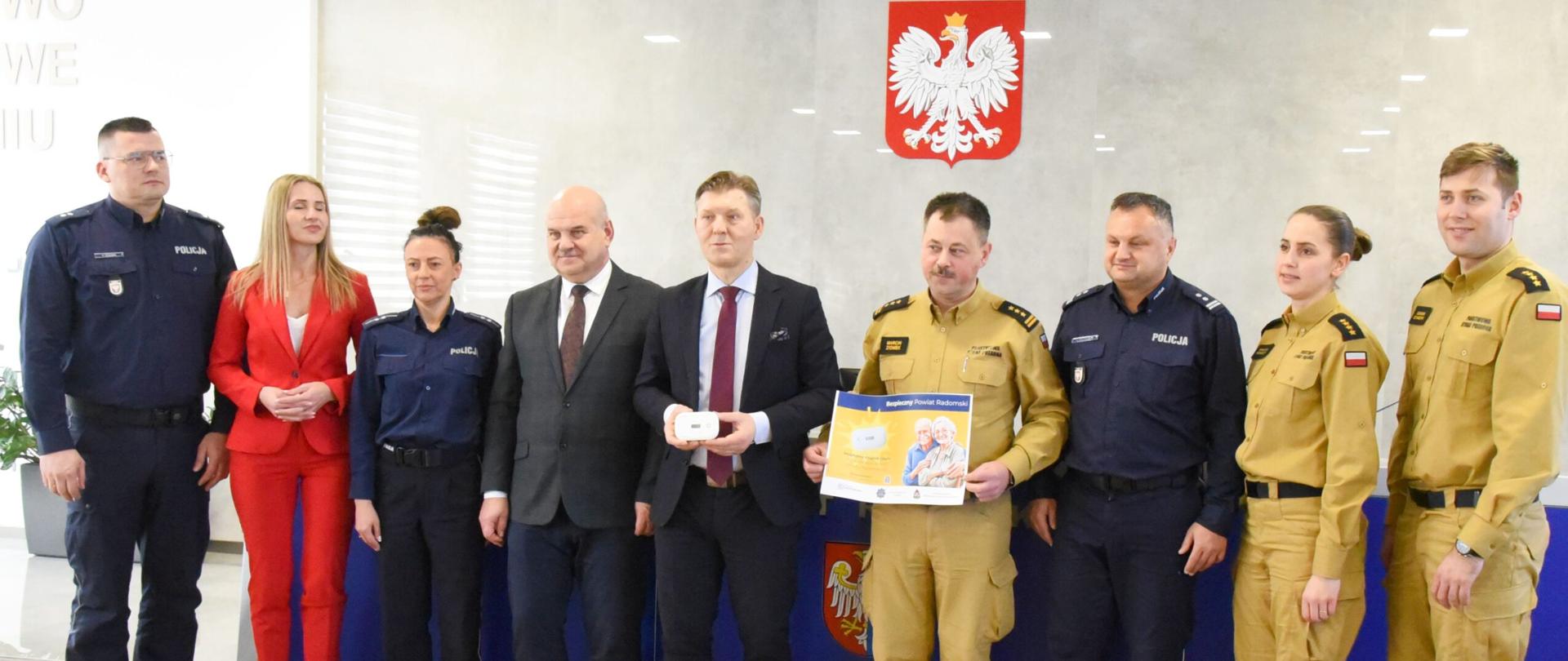 Zdjęcie wykonane w sali konferencyjnej w budynku starostwa powiatowego w Radomiu. Na zdjęciu widać stojące osoby pozujące do pamiątkowego zdjęcia: starostę radomskiego wraz z pracownikami starostwa, funkcjonariuszy Państwowej Straży Pożarnej oraz Policji. Starosta w rękach trzyma czujkę czadu. obok niego stoi zastępca komendanta miejskiego Państwowej Straży Pożarnej w Radomiu trzymający w rękach plakat promujący akcję babciu, dziadku nie czujesz - trujący, bezwonny, bezbarwny. 
