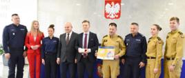 Zdjęcie wykonane w sali konferencyjnej w budynku starostwa powiatowego w Radomiu. Na zdjęciu widać stojące osoby pozujące do pamiątkowego zdjęcia: starostę radomskiego wraz z pracownikami starostwa, funkcjonariuszy Państwowej Straży Pożarnej oraz Policji. Starosta w rękach trzyma czujkę czadu. obok niego stoi zastępca komendanta miejskiego Państwowej Straży Pożarnej w Radomiu trzymający w rękach plakat promujący akcję babciu, dziadku nie czujesz - trujący, bezwonny, bezbarwny. 