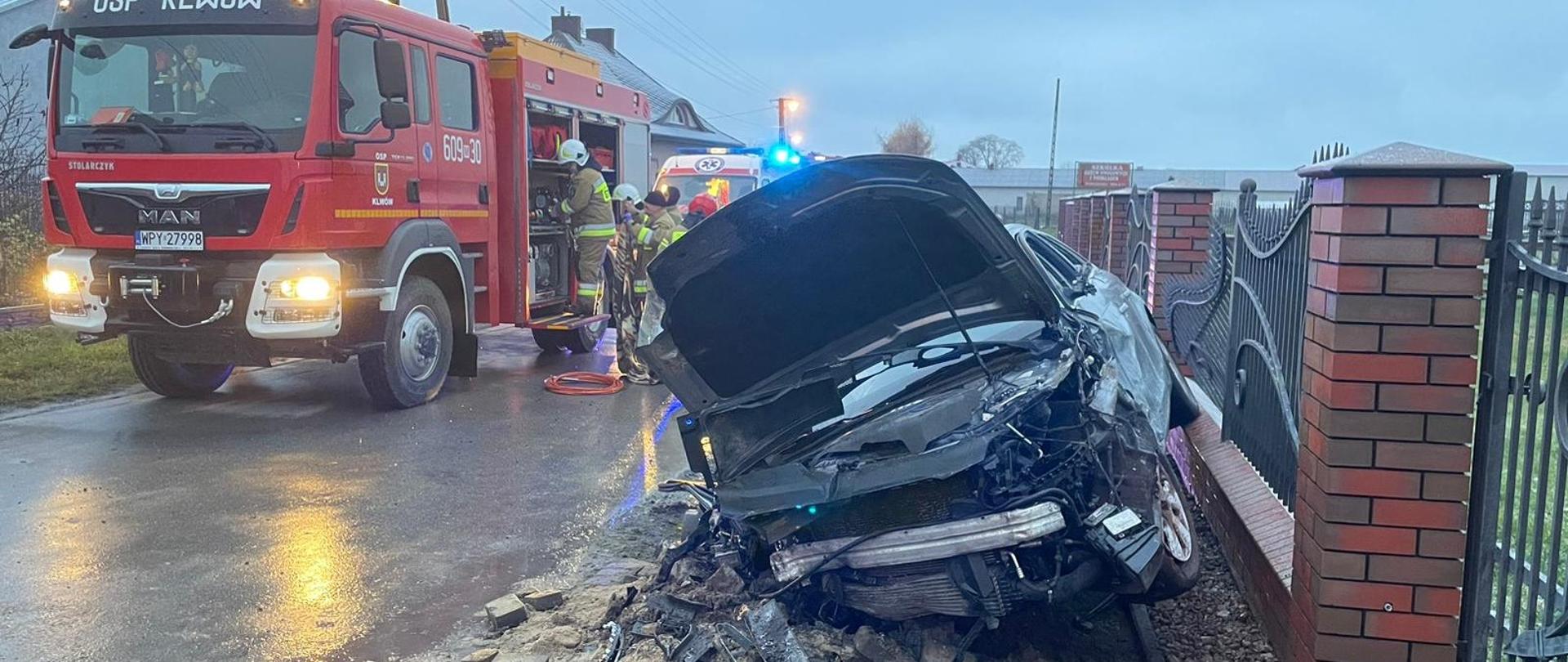 Zdjęcie przedstawia scenę wypadku drogowego, gdzie ciemny samochód osobowy uderzył w ceglany słup ogrodzenia, powodując znaczne zniszczenia. Jest to pochmurny dzień, a droga i teren są mokre, co wskazuje na niedawne opady deszczu. Na pierwszym planie, po prawej stronie, widać rozbity przód samochodu, który jest mocno zdeformowany. Elementy karoserii i silnika są rozrzucone na ziemi, a słup ogrodzenia jest częściowo zniszczony. Po lewej stronie kadru znajduje się czerwony wóz strażacki z napisem "OSP KLWÓW" i numerem "609-30". Obok wozu widać strażaków w żółtych kombinezonach oraz personel medyczny w białych kombinezonach z zielonymi odblaskami, co sugeruje trwającą akcję ratunkową. W tle, za wozem strażackim, widać niebieskie światła pojazdu uprzywilejowanego, prawdopodobnie karetki. Cała scena wskazuje na poważny wypadek drogowy z udziałem służb ratunkowych.