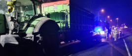 Uderzenie samochodu osobowego w przyczepę rolniczą. Droga wojewódzka nr 309. Na jezdni znajduje się ciągnik rolniczy koloru zielonego z przyczepą, za którą widoczny jest samochód osobowy. Obok pojazdów są strażacy uczestniczący w akcji. W tle rondo i lampy uliczne.