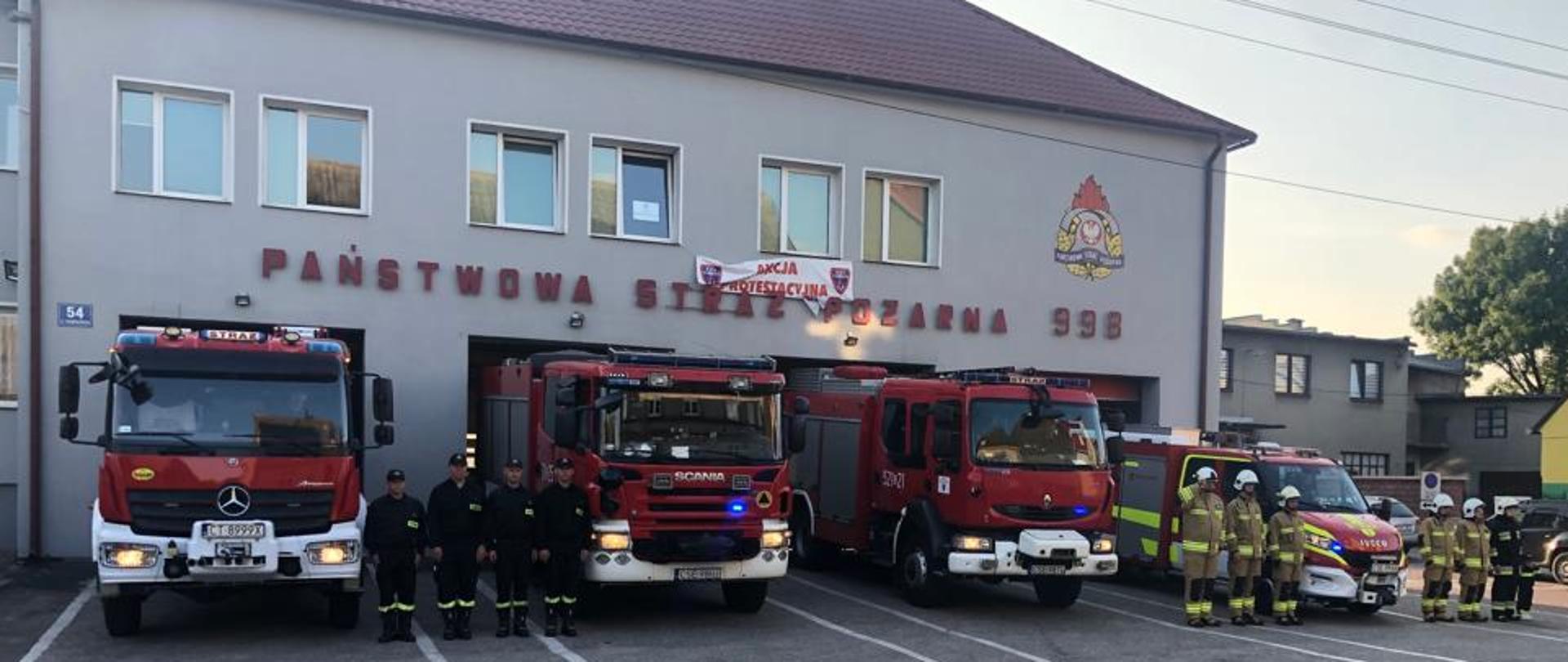 Strażacy Państwowej Straży Pożarnej ubrani w mundury koszarowe oraz strażacy Ochotniczej Straży Pożarnej ubrani w mundury specjalne stoją przed budynkiem komendy, obok nich stoją samochody ratowniczo gaśnicze.