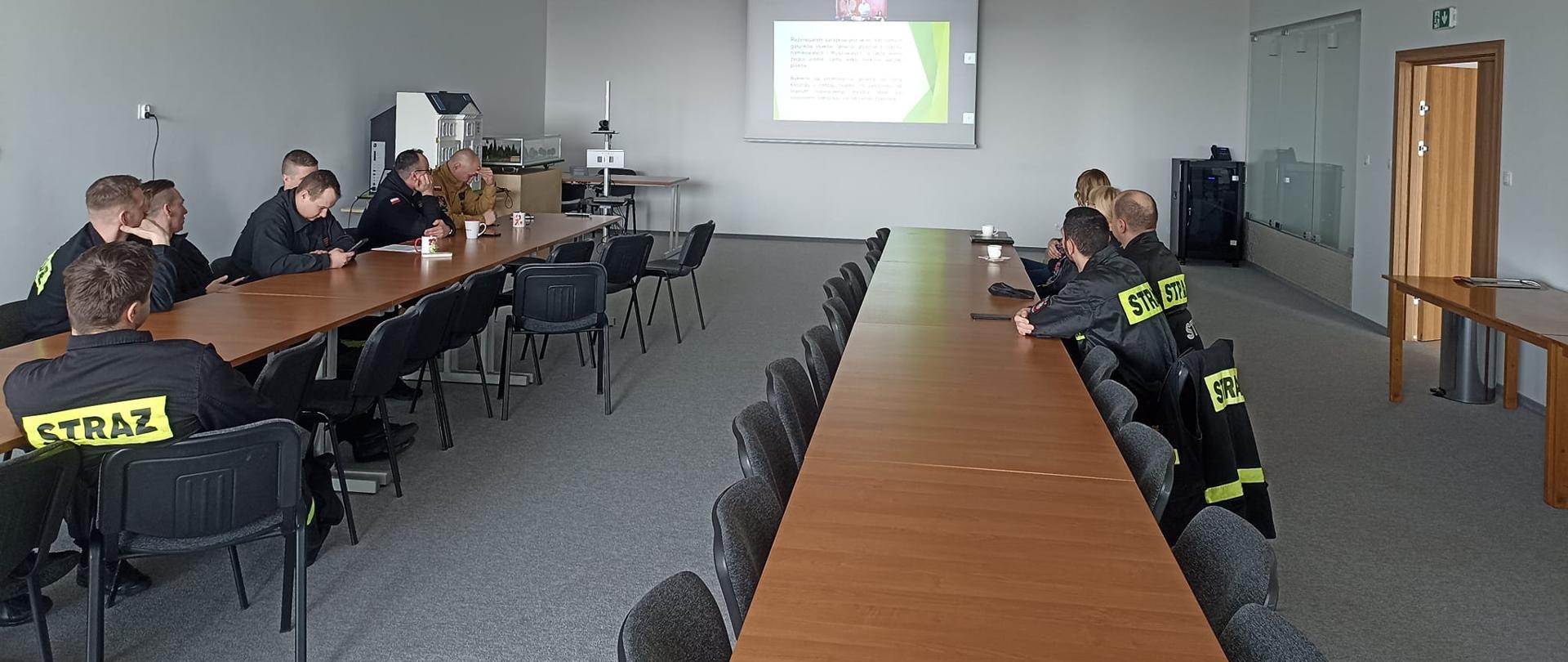 Zdjecie przedstawia strażaków uczestniczących w videokonferencji. Strażacy siedzą przy długich stołach