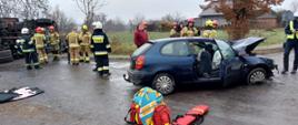 Strażacy oraz osoby uczestniczące w zdarzeniu znajdują się przy pojazdach. Na pierwszym planie sprzęt medyczny, którym udzielano pierwszej pomocy. W tle zabudowania.