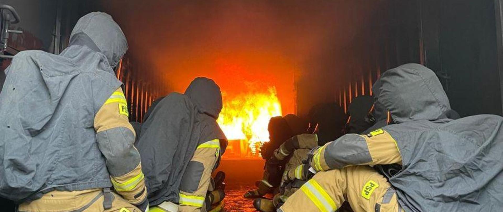 Strażacy Państwowej Straży Pożarnej w umundurowaniu bojowym siedzą w komorze rozgorzeniowej na poligonie ratowniczym Komendy Wojewódzkiej PSP w Opolu podczas szkolenia. W tle widoczne płomienie.