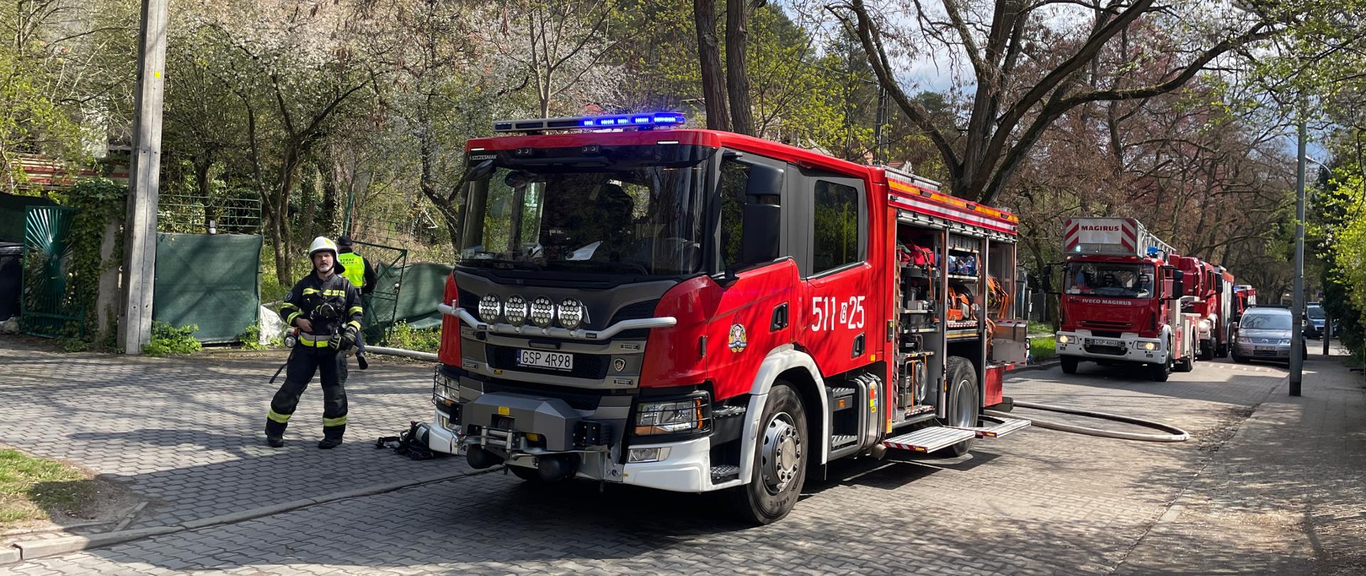 Samochody strażackie stoją na ulicy jeden za drugim. Pierwszy stoi GCBA Scanmia, za nim drabina mechaniczna SCD i dalej dwa kolejne prawie niewidoczne. Obok pierwszeg przechodzi strażak OSP
