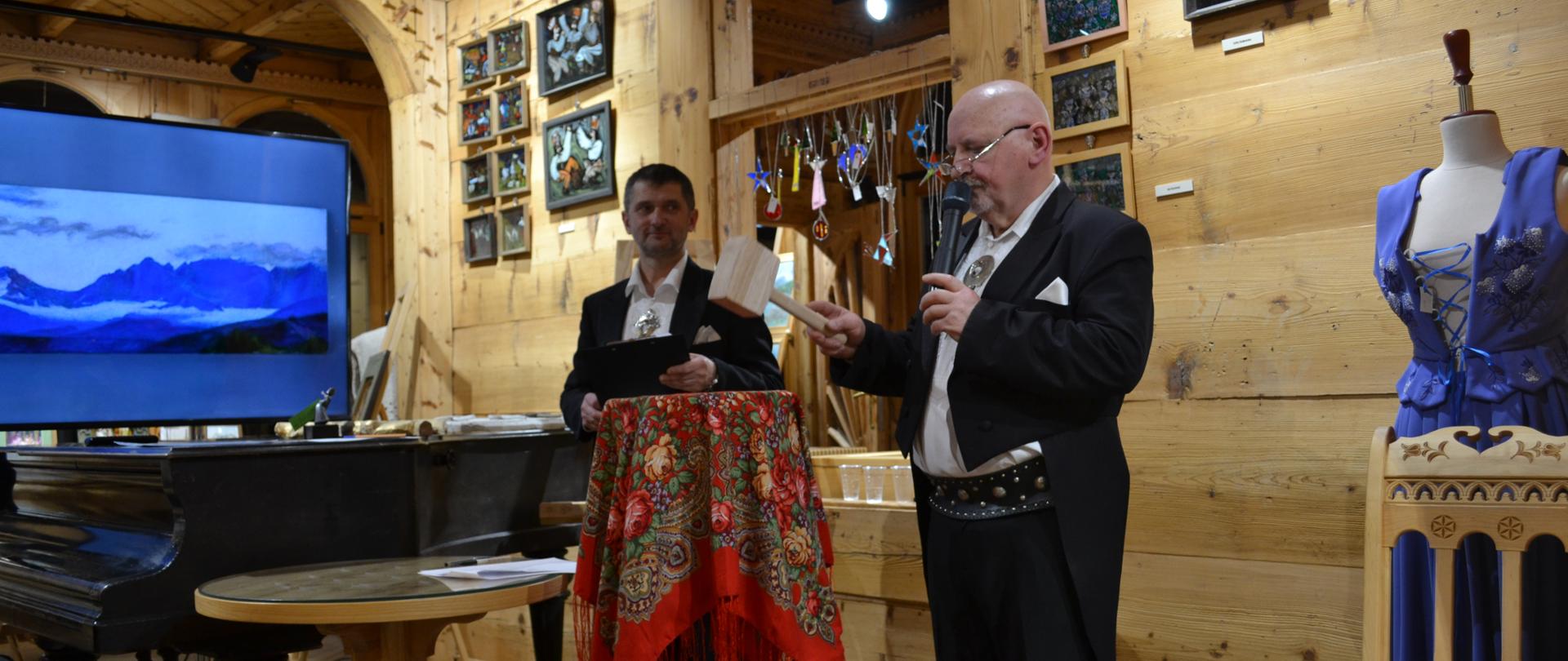 Na zdjęciu wnętrza zabytkowej willi "Czerwony Dwór" mieszczącej się przy ul. Kasprusie w Zakopanem, w której dnia 17 grudnia 2023 roku miała miejsce aukcja zorganizowana przez uczniów PLSP im. Antoniego Kenara w Zakopanem na rzecz budowy Pomnika Kurierów Tatrzańskich. Na pierwszym planie widzimy dwie postacie prowadzące licytację prac, są to mężczyźni ubrani w czarne smokingi i białe góralskie koszule. Do koszul mają dodatkowo przypięte góralskie, metalowe spinki góralskie. Po lewej stronie występuje pan Mirosław Spera a po prawej pan Tomasz Zieliński. Pan Tomasz w ręku trzyma drewniany młotek, którym uderza w drewniany kawalet pełniący rolę stolika przykrytego góralską chustką. Uderzenie młotka w blat stolika świadczy o zatwierdzeniu podanej kwoty licytowanego dzieła. 