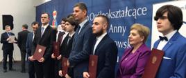 Zdjęcie z boku, wiceminister Piontkowski, kobieta ubrana na fioletowo i kilkunastu młodych ludzi w garniturach trzymających dyplomy w brązowych okładkach stoją w rzędzie pod granatową ścianą.