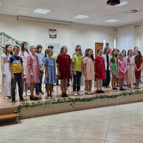 Chór szkolny podczas występu. Grupa dzieci w strojach galowych śpiewa, stojąc na podwyższeniu na scenie. Przed nimi stoi ubrana na czarno młoda dyrygentka.