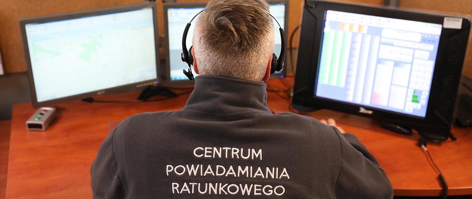 operator siedzi w słuchawkach przed trzema monitorami, na plecach ma napis"CENTRUM POWIADAMIANIA RATUNKOWEGO"
