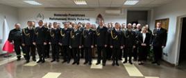 Na sali KP PSP Mława uroczysta zbiórka, zebrani strażacy pozują do zdjęcia grupowego.
