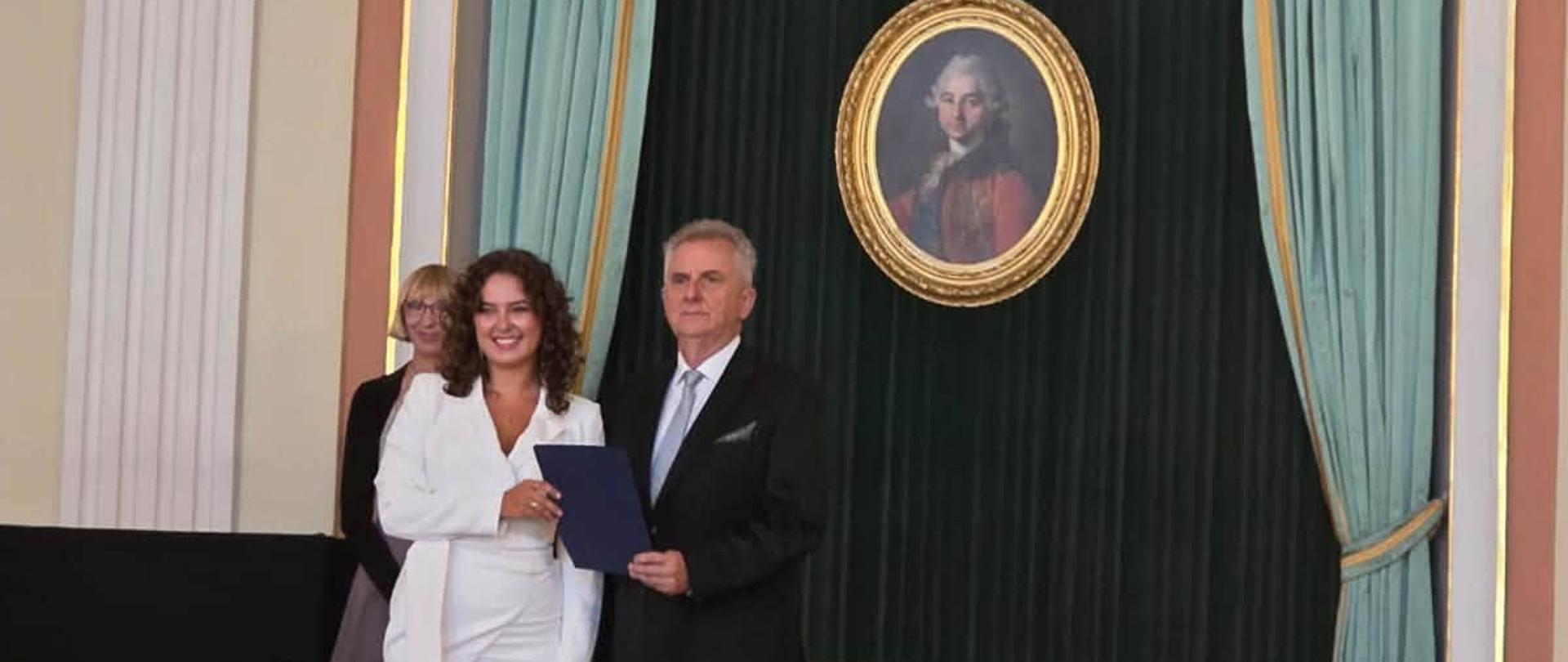 Kobieta w białej sukience i mężczyzna w garniturze. Z tyłu zabytkowy obraz. Widoczna publiczność.