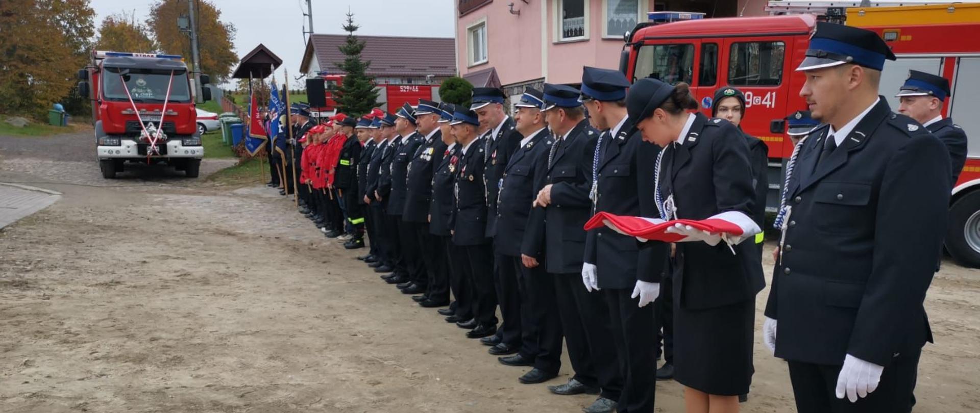 Druhowie Ochotniczej Straży Pożarnej w Pogódkach stoją w rzędzie przed samochodem gaśniczym.