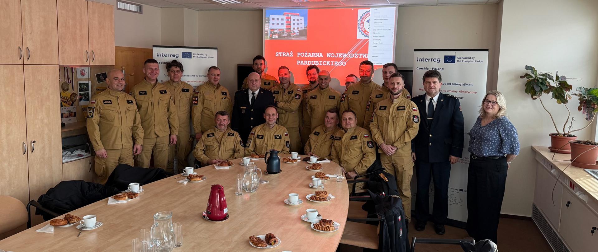Zdjęcie grupowe zrobione w sali konferencyjnej. Około dwudziestu osób, w większości strażaków w beżowych mundurach, oraz dwie osoby w strojach cywilnych/galowych, pozują do zdjęcia za długim stołem. Na ścianie za nimi wyświetlana jest prezentacja z napisem "STRAŻ POŻARNA WOJEWÓDZTWA PARDUBICKIEGO" i logo projektu Interreg. Na stole leżą filiżanki z kawą i drożdżówki