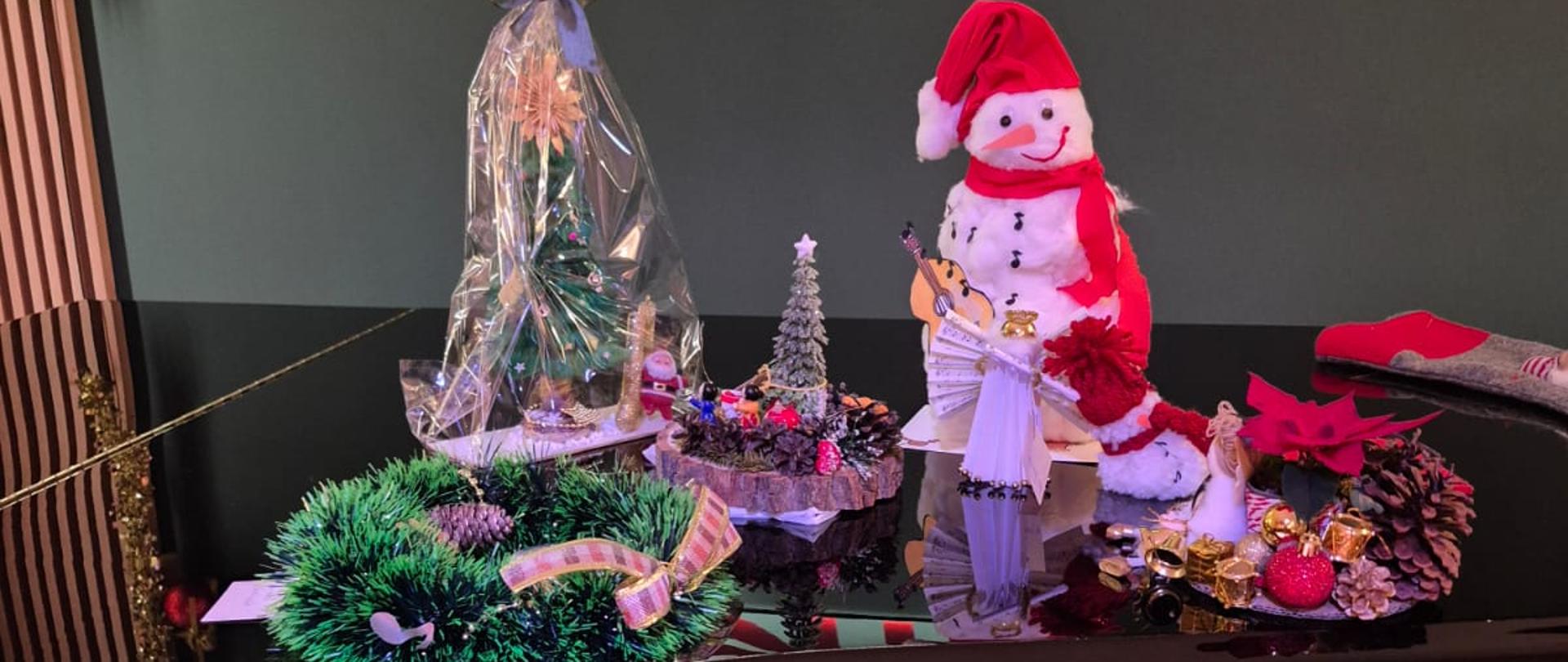 Świąteczna dekoracja umieszczona na fortepianie. Na blacie widoczne są: ozdobna, duża nutka z czerwoną czapeczką, figurka bałwanka w czerwonym stroju trzymającego nuty, a także ozdobne opakowanie z małą choinką i innymi dekoracjami. W tle widać ozdobione ściany z girlandami, bombkami oraz logo z literami 'PSM'. Po lewej stronie znajduje się oświetlona choinka