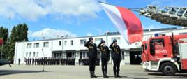 Trzech strażaków w galowych mundurach salutuje przed dużą flagą Polski zawieszoną na drabinie wozu strażackiego podczas uroczystości na placu jednostki PSP. W tle pozostali strażacy stoją w szeregu przed budynkiem strażnicy.