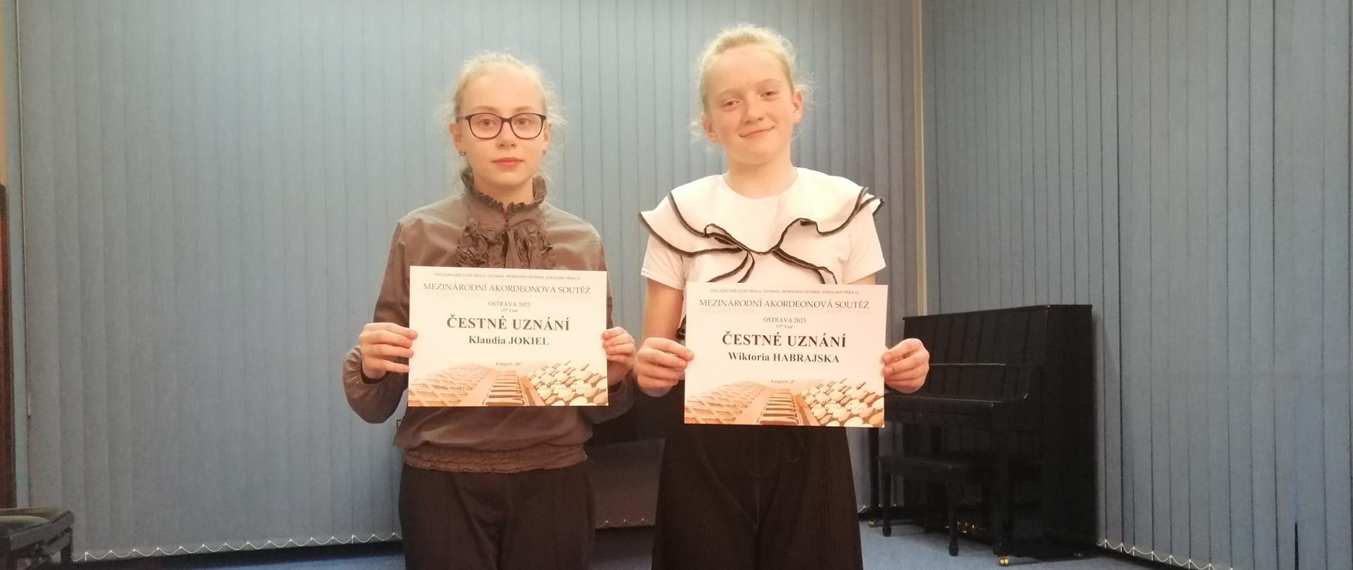 Klaudia Jokiel i Wiktoria Habrajska - uczennice klasy akordeonu p. Sławomira Mocha - z dyplomami na tle rolet w kolorze jasnoniebieskim. Z prawej strony w tle stoi czarne pianino. 