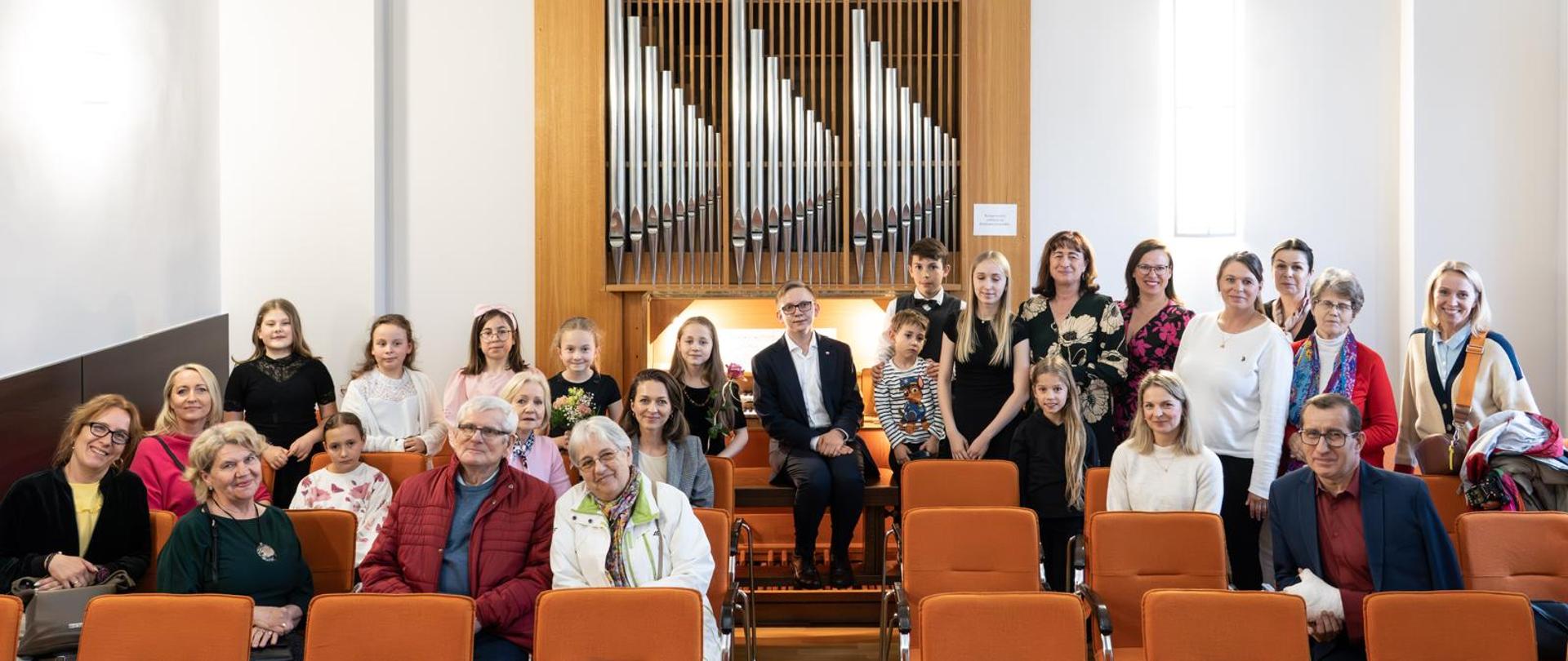 Na zdjęciu uczniowie odświętnie ubrani. Po prawej stronie zdjęcia kobieta brunetka w czarnej sukience w beżowe kwiaty. Przed nimi pomarańczowe krzesła, a na nich zgromadzona publiczność- rodzice i dziadkowie uczniów. W tle za nimi piszczałki organowe.