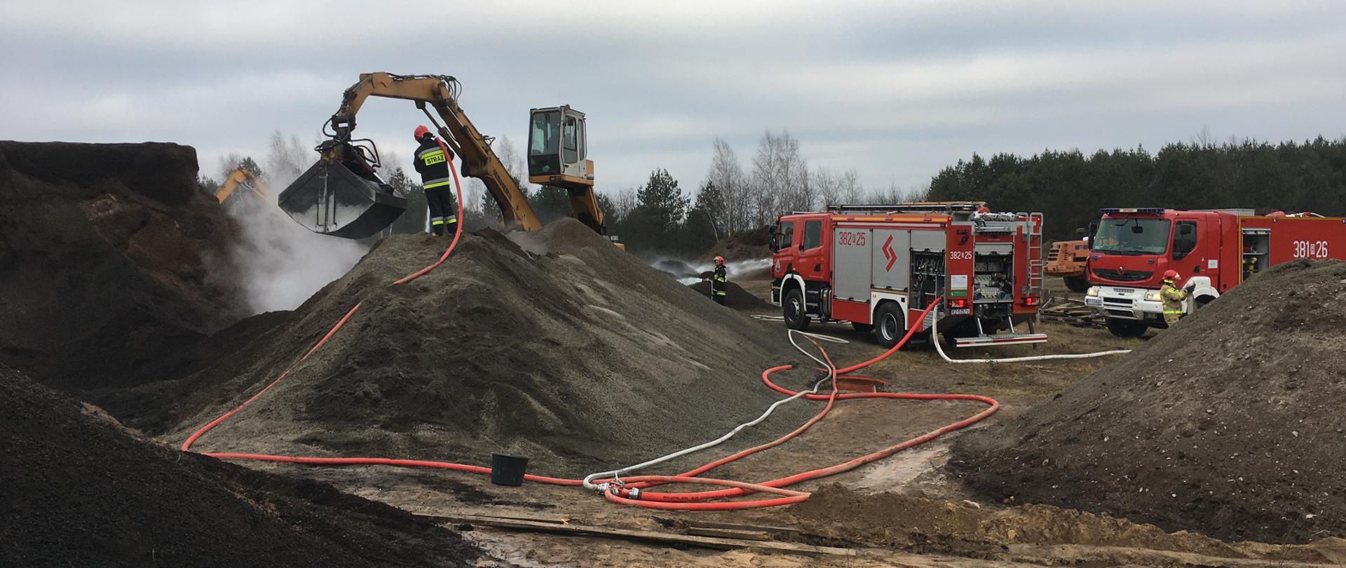 Teren akcji gaśniczej. Koparka przesypuje palący się pellet, a strażak stojący na hałdzie przelewa materiał wodą. Na placu zgromadzona jest duża ilość węży gaśniczych, które zasilane są przez dwa samochody gaśnicze. Z wnętrza hałdy wydobywa się dym. 