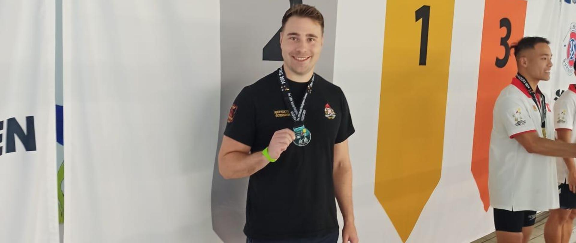 Na zdjęciu widać trzech mężczyzn z medalami zawieszonymi na szyi. Jeden pozuje do zdjęcia a dwaj stoją obok. Ubrani są w koszulki na krótki rękawek oraz krótkie spodenki. W tle widać baner z napisem SVOM DANMARK oraz logo i flagami Danii oraz numerami podium. Zdjęcie zrobione jest na krytym basenie.