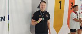 Na zdjęciu widać trzech mężczyzn z medalami zawieszonymi na szyi. Jeden pozuje do zdjęcia a dwaj stoją obok. Ubrani są w koszulki na krótki rękawek oraz krótkie spodenki. W tle widać baner z napisem SVOM DANMARK oraz logo i flagami Danii oraz numerami podium. Zdjęcie zrobione jest na krytym basenie.