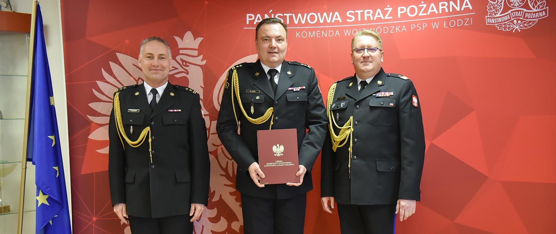 brygadier Lusa brygadier Łukasik oraz nadbrygadier Janowski pozują do zdjęcia na tle baneru komendy wojewódzkiej,