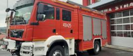 Fotografia przedstawia czerwony, nowy, ciężki samochód ratowniczo-gaśniczy na tle budynku remizy Komendy Powiatowej Państwowej Straży Pożarnej w Brzozowie. Na fotografii widać bojowy samochód pożarniczy, jego przód i lewy bok. 