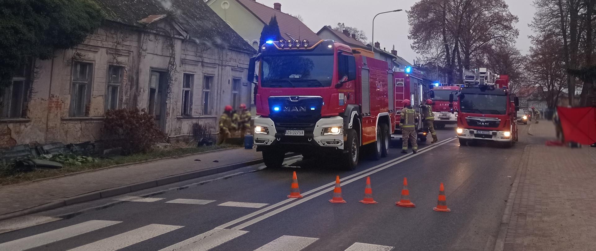 Na zdjęciu na pierwszym planie znajdują się wozy strażackie ustawione na drodze. Pojazdy zajmują dwa pasy ruchu blokując ruch pojazdów i zabezpieczając teren akcji. W tle widoczny budynek typu dom jednorodzinny. Wokół pojazdów strażacy wyciągający sprzęt niezbędny do działań.