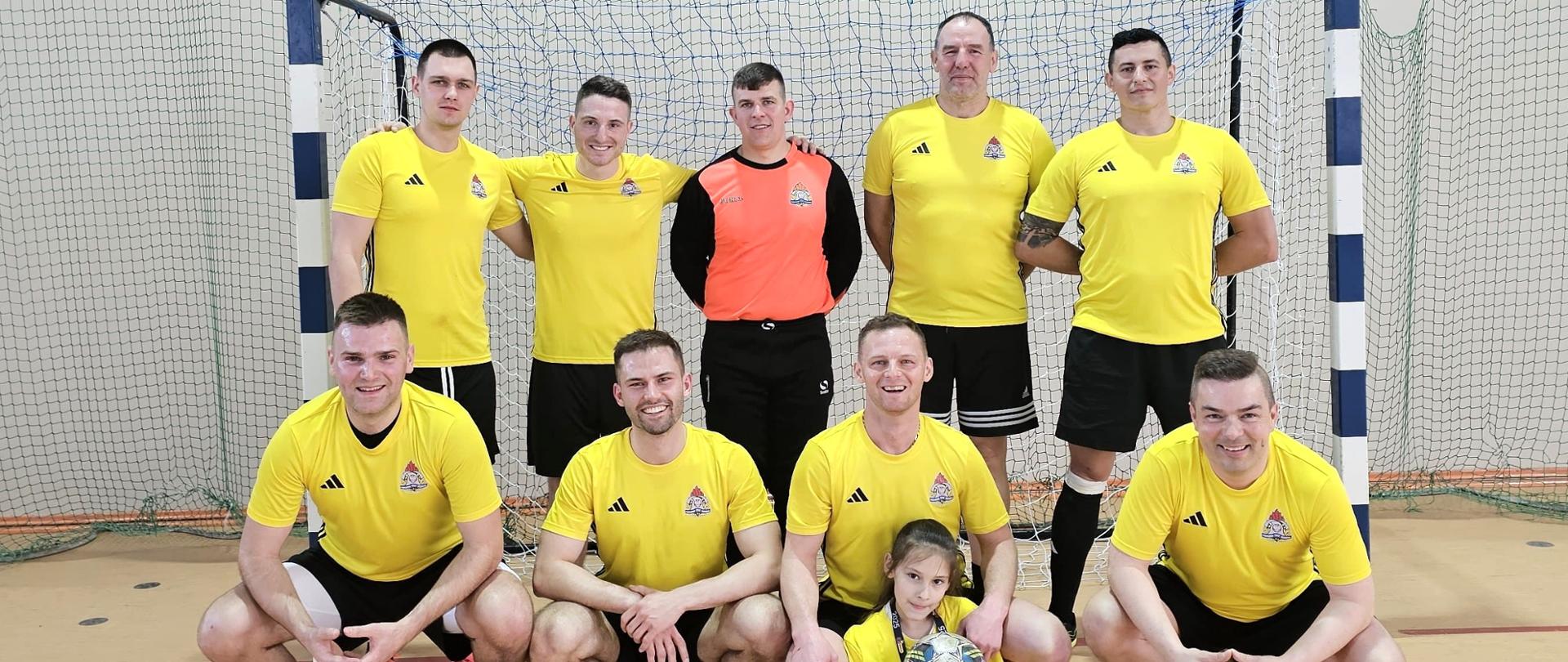Na zdjęciu została przedstawiona drużyna KP PSP w Kartuzach. Strażacy ubrani w strój sportowy stoją na tle bramki.