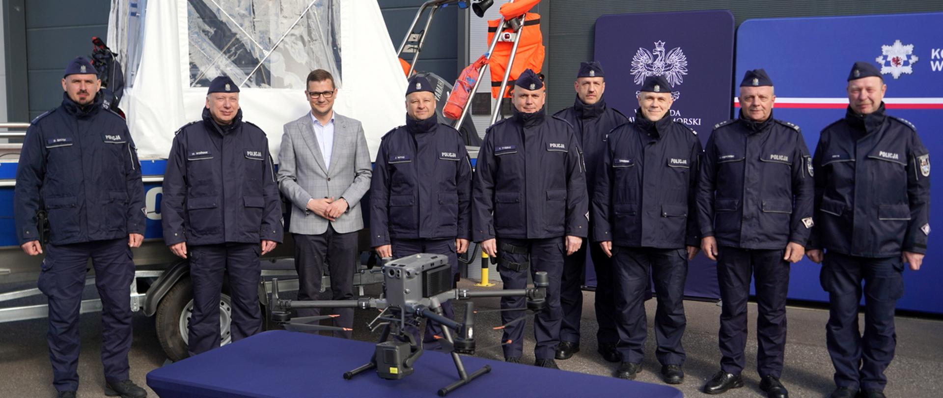 Wojewoda Kujawsko-Pomorski Michał Sztybel stoi między policjatami. Obok niego stoją policjanci, a za nimi stoi ścianka reklamowa komendy wojewódzkiej policji, zakupiony sprzęt oraz rollup z napisem wojewoda kujawsko-pomorski. 