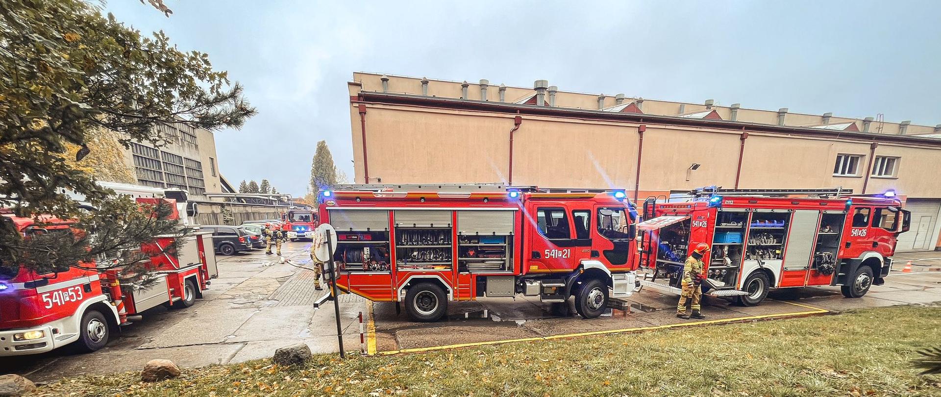 Samochody strażackie stoją przy budynku szkoły. Na drugim planie widać zaparkowane samochody oraz strażaków .
