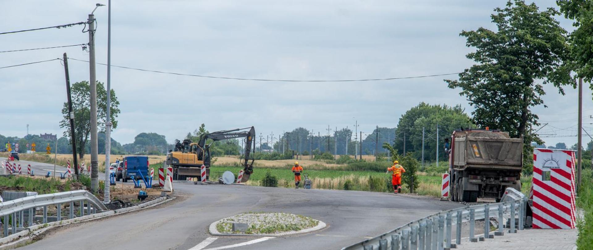 Roboty drogowe na odcinku wiejskiej drogi: koparka, samochód ciężarowy i pracownicy w odzieży ostrzegawczej naprawiają nawierzchnię; jezdnia jest częściowo zamknięta, ustawiono barierki i oznakowanie, w tle pola i linia energetyczna.