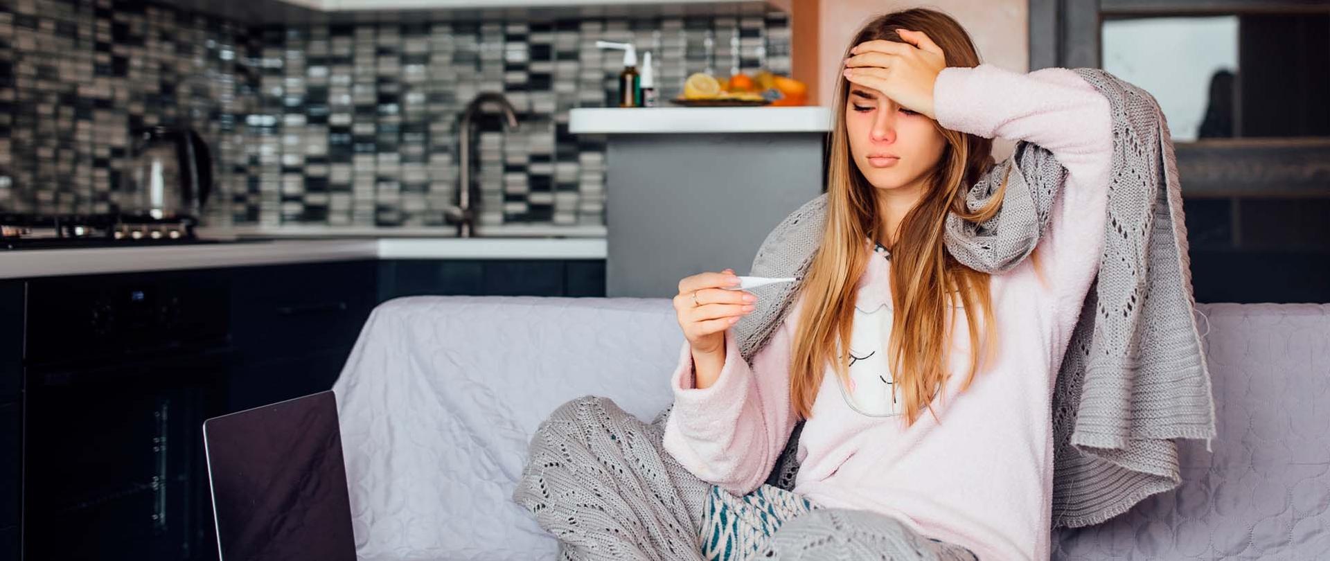 Chora kobieta siedzi na kanapie owinięta kocem z termometrem w ręku.