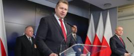 Zdjęcie z boku - przy dwóch mównicach z przezroczystego tworzywa stoją minister Czarnek który mówi do mikrofonu i mężczyzna w garniturze, za nimi trzech mężczyzn, za nimi dalej ściana, po bokach z obu stron polskie flagi.