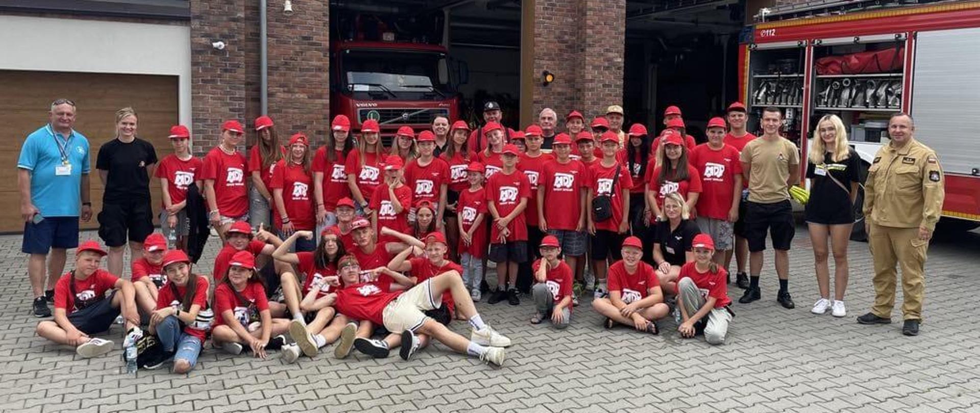 Na zdjęciu uczestnicy obozu wraz z opiekunami. Po prawej stronie stoi Komendant Powiatowy PSP w Pucku Pan bryg. Wojciech Wanat. Komendant jest ubrany w ubranie służbowe w kolorze piaskowym. Na zdjęciu również funkcjonariusze st.sekc. Aleksandra Dettlaff-Rambiert oraz st.str. Wojciech Stachowicz ubrani w ubranie koszarowe. Młodzież ma ubrane czerwone koszulki oraz czerwone czapki z napisem ,,MDP''