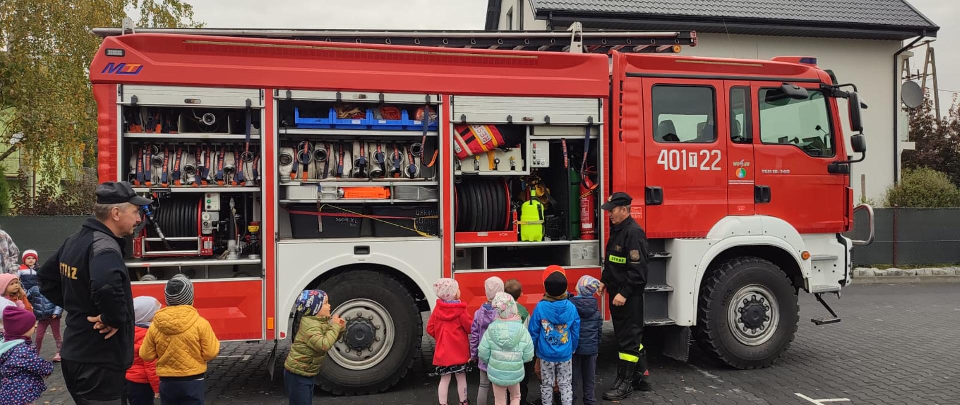 Na 1 planie przedszkolaki i 1 strażak, przed dziećmi stoi strażak oraz samochód strażacki z otwartymi skrytkami. 