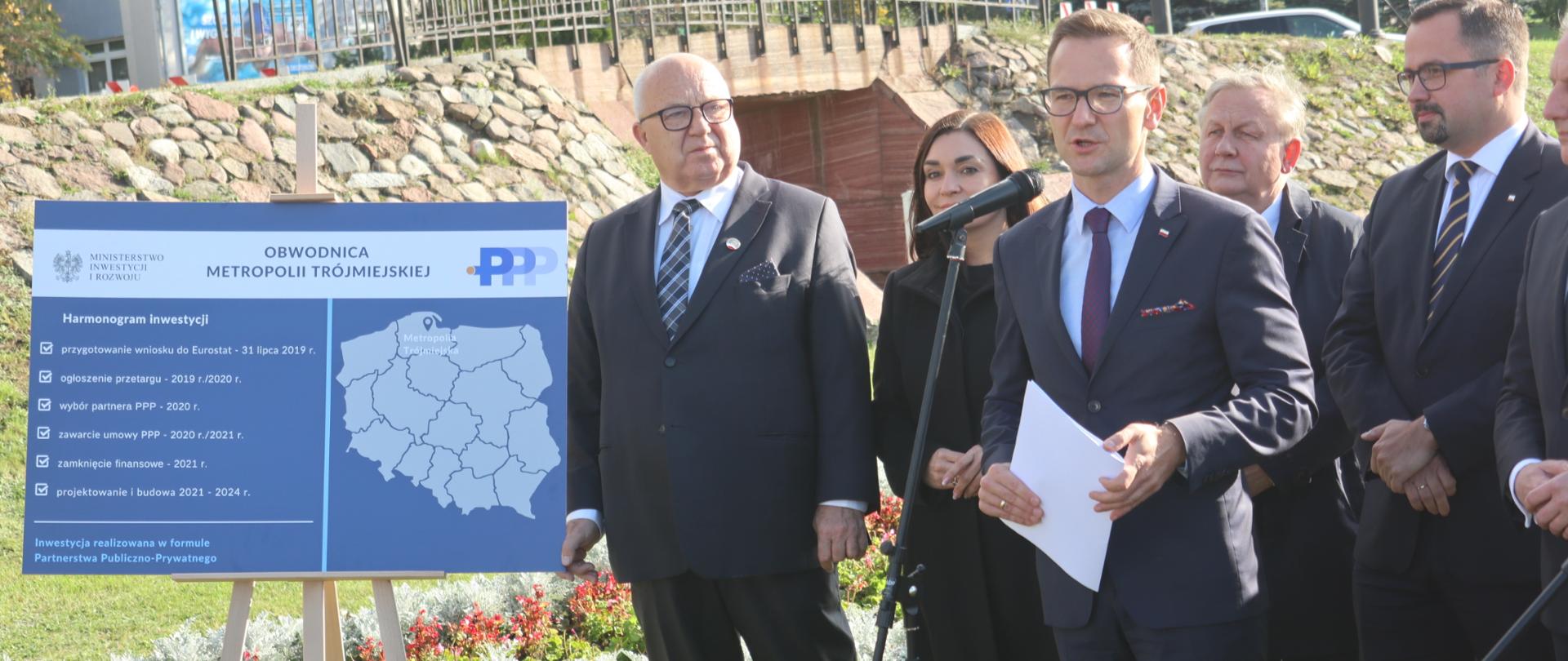 Na zdjęciu stoi pięć osób, wiceminister Waldemar Buda trzeci z lewej, skrajnie po lewej stronie widać tablicę z harmonogramem budowy.