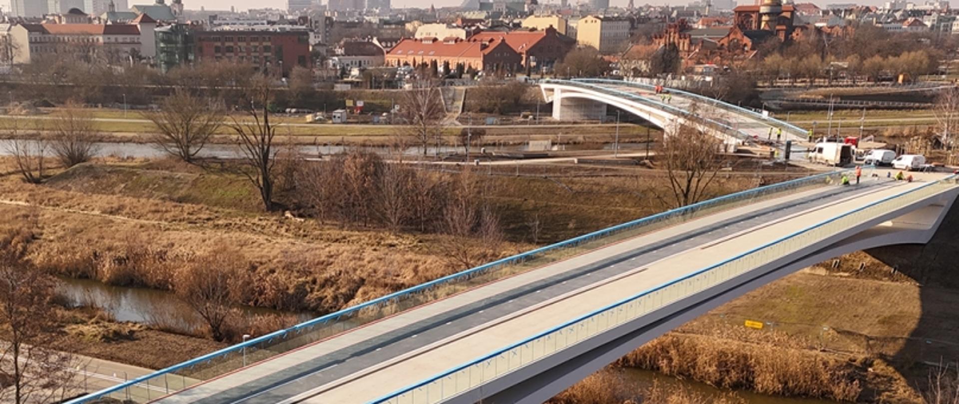 Na zdjęciu widać dwie kładki pieszo-rowerowe z lotu ptaka. W oddali widać zabudowania.