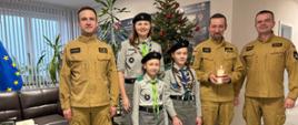 Trzech funkcjonariuszy komendy w strojach służbowych oraz trzy harcerki, jedna trzyma lampion ze świecą. Wszyscy stoją na tle choinki w holu jednostki 
