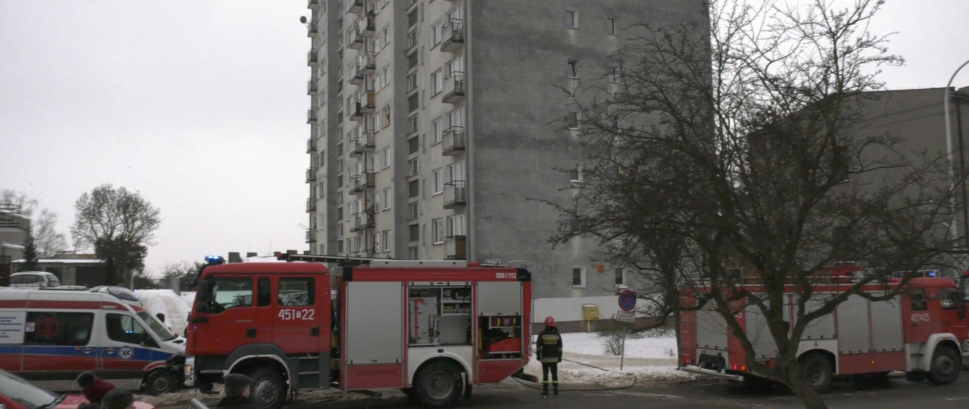 Na pierwszym planie samochody strażackie i karetka pogotowia, w tle budynek mieszkalny wielorodzinny w którym doszło do pożaru.