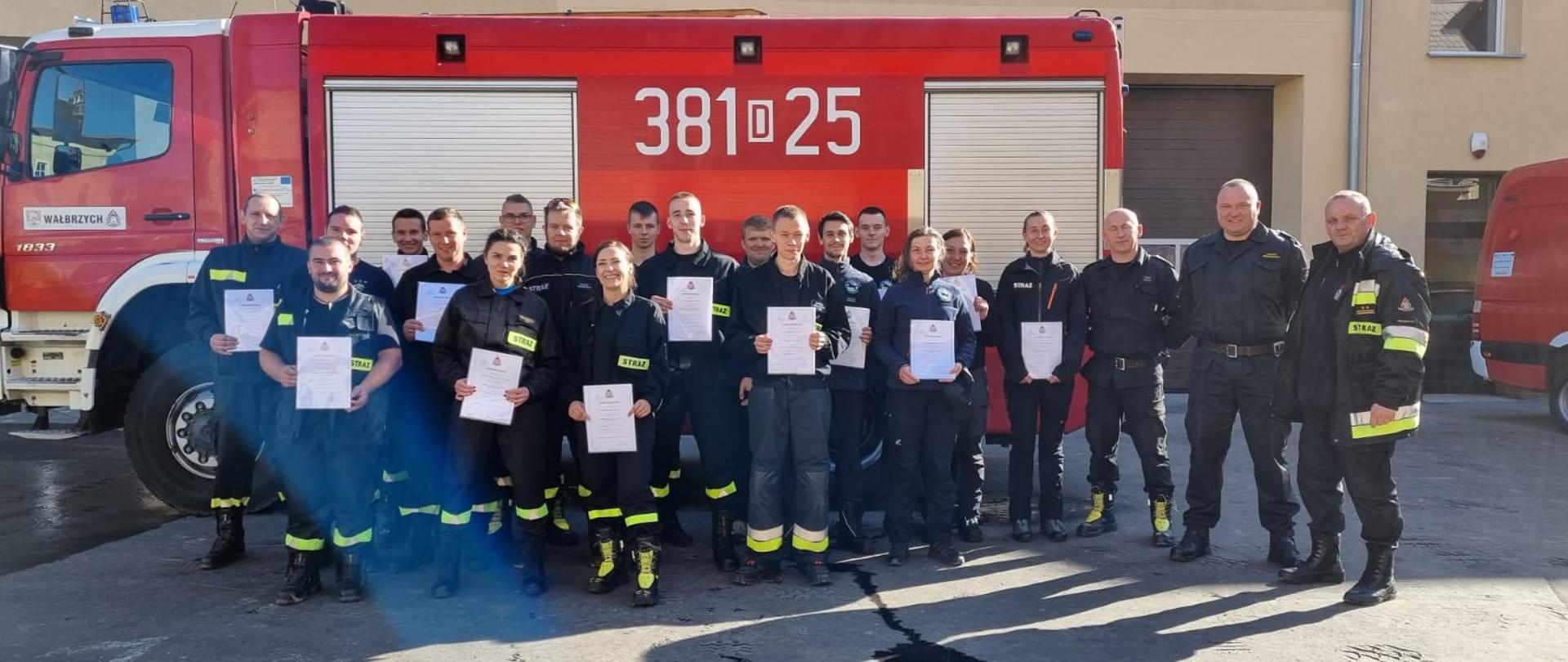 Strażacy stoją na placu JRG nr 1 w Wałbrzychu po zdanym egzaminie, trzymają w rękach dyplomy. W tle czerwony wóz strażacki i budynek